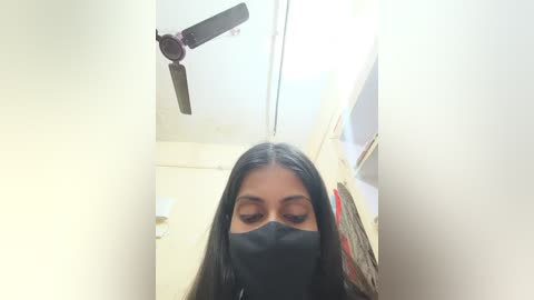 A video of a South Asian woman with long black hair, wearing a black face mask, standing in a dimly lit, beige-tiled bathroom. A ceiling fan hangs above.
