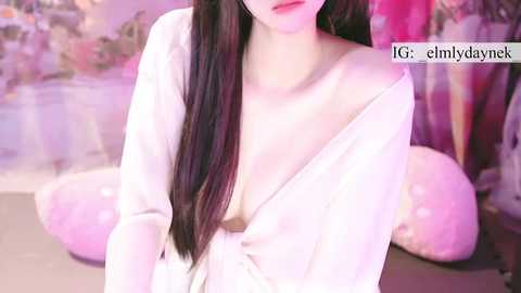 Video of a light-skinned woman with long dark hair, wearing a sheer white dress that reveals her breasts, kneeling on a soft surface. Background features pastel-colored, abstract art and soft lighting.