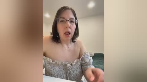 A video of a woman with shoulder-length brown hair, wearing glasses, a gray off-shoulder top, and a necklace, in a modern bedroom with beige walls, a green bed, and recessed ceiling lights.