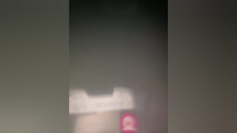 A blurred video featuring a woman's face partially obscured by a black vertical object, likely a curtain or a screen. The background is dark and indistinct, with a pinkish light source below.