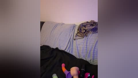 A video of a child's bedroom with a messy bed featuring a light blue blanket, a black and white patterned pillow, and a stuffed toy. The background is a plain white wall, and the scene appears dimly lit.