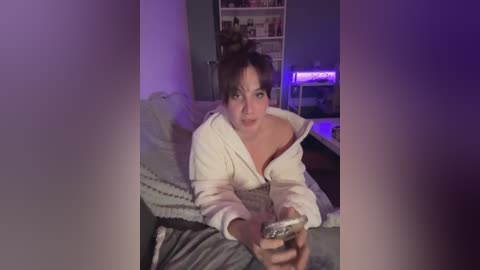 Video of a woman with dark hair, wearing a white robe, sitting on a bed with a grey blanket, holding a remote control. Background features a bookshelf and dim lighting.