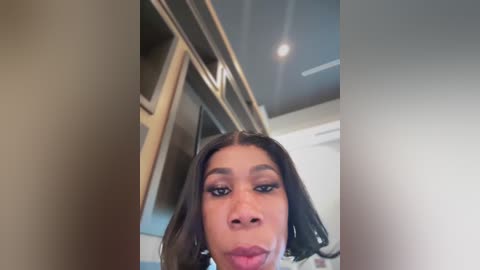 A candid, close-up video of a woman with medium brown skin and straight, shoulder-length dark hair, wearing subtle makeup and a neutral expression. Background features a modern, minimalist room with neutral tones and a sleek, white ceiling light.