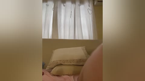 Video of a bedroom with beige walls, featuring a bed with a beige pillow and white curtains slightly open, revealing a glimpse of the window and a small part of a person's bare torso.