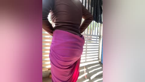 Media: Video of a person, likely female, with a dark complexion, wearing a purple sari and a black long-sleeved top, standing indoors near a window with vertical blinds, casting striped shadows on the wall.
