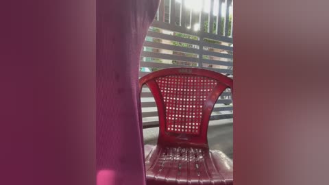 Video of a red plastic chair with a lattice backrest, placed in an outdoor setting with a metallic fence and greenery in the background. The image is taken from an angle, creating a sense of depth.