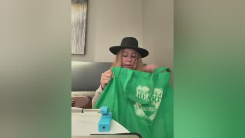 A video of a blonde woman wearing a black hat and green apron, holding a green apron with \"Piggy Bank\" text. She's seated at a table with blue piggy banks and a white surface, in a minimalist room with beige walls and abstract art.