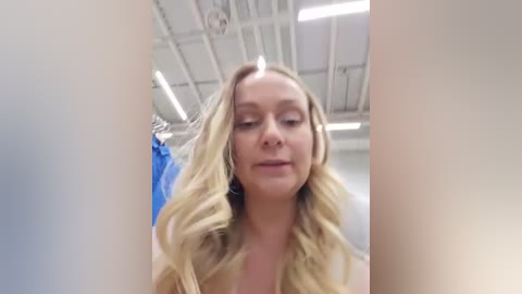 Video of a blonde woman with wavy hair, wearing a blue top, in an industrial setting with white ceilings and fluorescent lights.