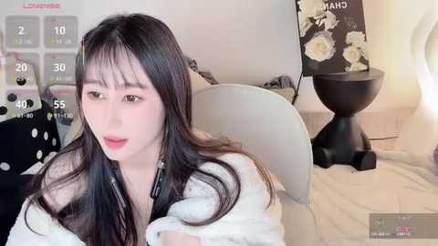 A video of a young East Asian woman with long, straight black hair, fair skin, and a slender physique, wearing a white robe, seated in a modern bedroom with a black lamp, white chair, and floral wallpaper.