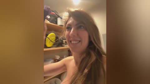 Video of a smiling, topless woman with long brown hair in a cluttered, dimly lit room. Shelves behind her hold various items, including a yellow object and a black backpack.