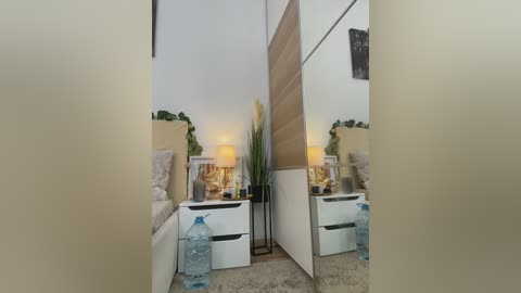 Video of a small, modern, minimalist bedroom with beige walls, a white bed, a wooden headboard, a table lamp, and a mirror reflecting the room's contents.