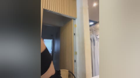 Video of a modern, minimalist bathroom with light grey walls, a striped wooden wall panel, and a beige towel rack. A person, partially visible, is wearing a black top and holding a cup, standing in front of a mirror.