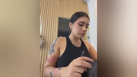 A video of a Latina woman with medium skin tone, dark hair in a bun, wearing a black tank top, and visible tattoos on her arms, holding a phone in a beige hallway with vertical wooden panels.