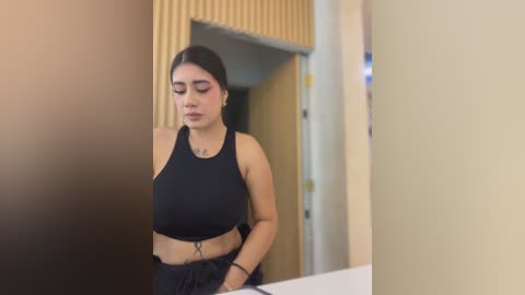 Video of a young Latina woman with medium skin tone, long black hair, and full lips, wearing a black sports bra and black pants, standing in a modern, beige-toned room with a wooden door.