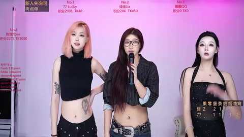 Video of three East Asian women, one with pink hair, one with straight black hair, and one with long dark hair, wearing casual attire, speaking into microphones against a purple and pink backdrop.