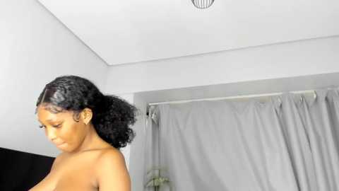 A video of a topless Black woman with curly hair, standing in a modern, minimally decorated room with white walls, grey curtains, and a ceiling fan.