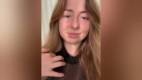 A video of a young woman with fair skin and long, wavy blonde hair. She has a soft, gentle smile and is wearing a black hairdresser's cape. The background is blurred, focusing attention on her face.