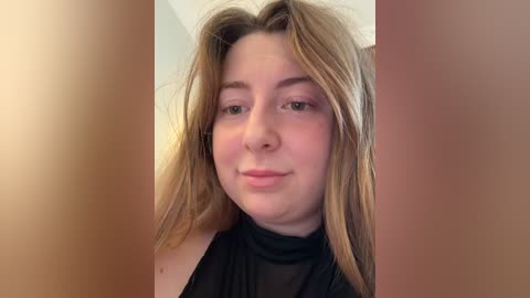 A close-up video of a young woman with light skin and long, straight blonde hair, wearing a black turtleneck. The background is blurred, highlighting her facial features.