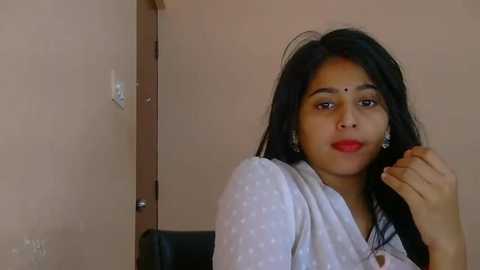 Video of a young woman with long black hair, wearing a white polka-dotted saree, sitting indoors against a beige wall. She has a serene expression, red bindi, and silver earrings.