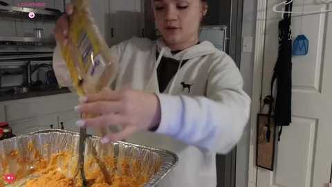 Video of a young boy with light skin and brown hair in a white hoodie, holding a plastic bag with yellowish-orange food. He's in a modern kitchen with white cabinets and a door.