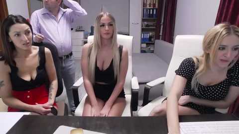 Video of four women in an office, three in revealing outfits, one in a polka-dot dress, white leather chairs, shelves, and bookcases in the background.