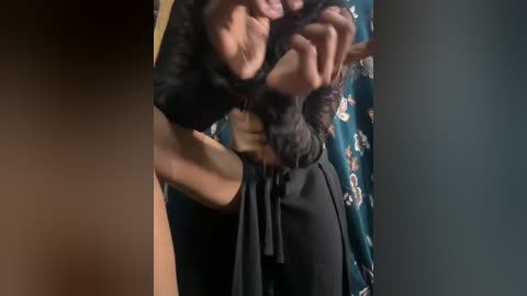 Video of a woman with medium skin tone, curly dark hair, and a black dress, playfully pulling the dress' tie with her teeth, in a dimly lit room with floral curtains.