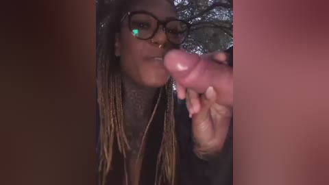 Video of a Black woman with dreadlocks and glasses, wearing a black shirt, holding a dildo to her mouth, taken in dim lighting with blurred background.