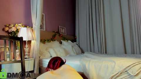 Video of a cozy, dimly lit bedroom with a white canopy bed, white bedding, beige curtains, and a red-haired child partially visible under the covers.