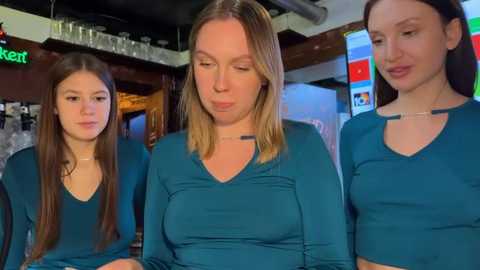 Video of three women in teal tops, two younger, one older, in a bar with glassware and TV screens in the background.