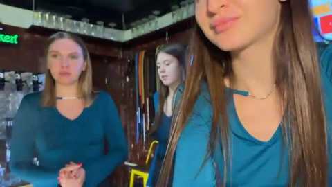 Video of three young women with long hair in teal long-sleeve shirts, standing in a dimly lit bar with wooden walls and hanging glasses.