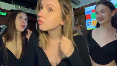 Video of three young women with light skin, wearing black tops, in a dimly-lit bar, with a colorful digital screen and liquor bottles in the background.