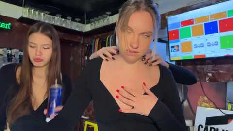 Video of two women, one blonde with a black top, massaging the other's chest in a dimly lit room with colorful TV screens and shelves.