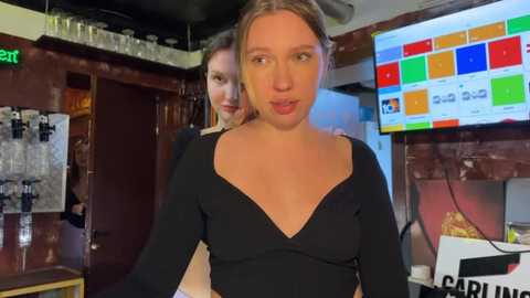 Video of a young woman with blonde hair in a black V-neck top, standing in a dimly lit bar with a colorful LED display and shelves in the background.
