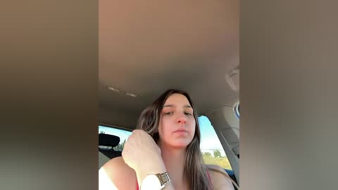 A video of a young woman with long brown hair, wearing a sleeveless top, sitting in a car with a blurred background, looking directly at the camera.