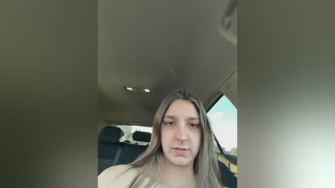 A video of a young woman with long, straight, brown hair, sitting in a car with dark upholstery, looking straight at the camera.