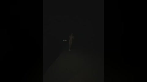 A dimly lit video of a lone figure walking on a deserted, dark street. The figure is indistinct and barely visible, creating a sense of mystery and isolation. The background is almost entirely black, emphasizing the eerie atmosphere.
