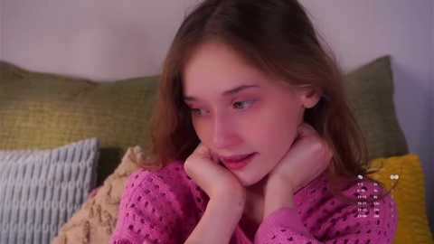 Video of a young woman with fair skin, long brown hair, wearing a pink knitted sweater, resting her chin on her hands, surrounded by textured cushions on a beige couch.