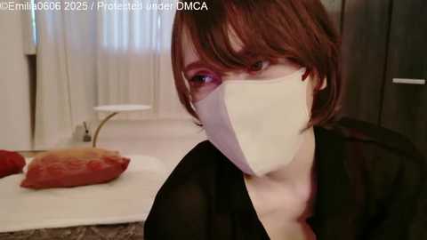 A video shows a person with short, auburn hair, wearing a white face mask and black clothing, standing indoors with white curtains and a table in the background.