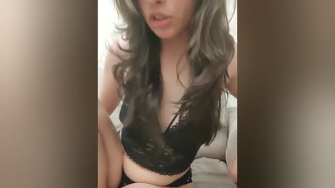 A video of a young woman with long, wavy, grayish hair, wearing a black lace bralette, sitting on a bed with beige sheets. The background is blurred, focusing on her partially exposed chest and lips.