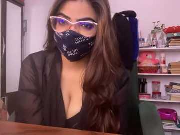 Video of a woman with long brown hair, wearing a black mask, glasses, and a sheer black robe, sitting at a cluttered desk in a messy office.
