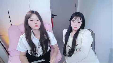 Video of two East Asian women with long black hair, fair skin, and red lipstick, dressed in white blouses and black skirts, sitting on pink chairs in a minimalist, white-walled room with a closed door.