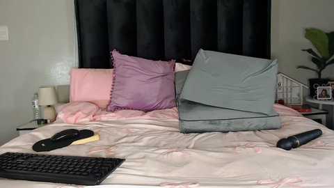 A video of a neatly made bed with a grey pillow and a pink pillow, black remote control, and a black keyboard on a white sheet.