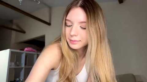 Video of a young Caucasian woman with long, blonde hair, wearing a white tank top, smiling softly, in a minimalist, well-lit room with beige walls and a white bookshelf in the background.