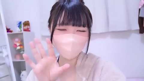 Video of an East Asian woman with black hair in a ponytail, wearing a face mask, waving with a soft, gentle expression. The background features a white room with a shelf holding toys and a pink item.