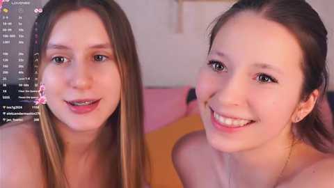 Video of two young women with light skin, smiling, wearing minimal makeup, with long hair, one with a pink flower in her hair. Background shows a pink and orange bed.