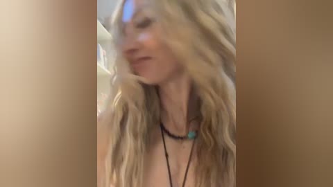 A blurry video of a young, fair-skinned woman with long, wavy blonde hair, wearing a black necklace with a turquoise pendant. She has a slender physique and is seen from the shoulders up, likely indoors.