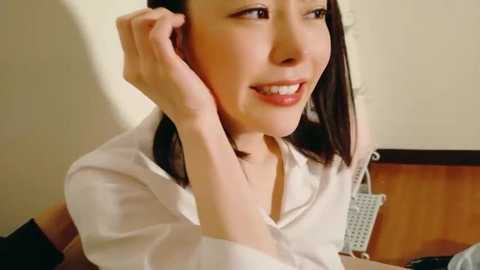 Video of an East Asian woman with straight, shoulder-length black hair, wearing a white blouse, smiling, resting her chin on her hand, in a softly lit room.