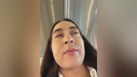 Video of a woman with long, dark hair, light brown skin, and a nose ring, winking and smiling, surrounded by blurred, metallic surfaces, possibly inside a car.