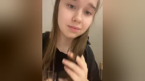 Video of a young girl with light skin and long, straight brown hair, slightly parted, holding a brush in her right hand. She wears a black shirt. Background is a beige wall and a wooden door.