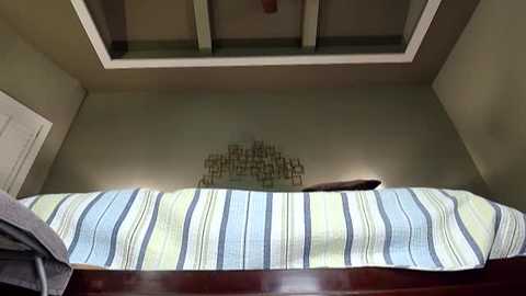 Video of a minimalist bedroom ceiling with a recessed skylight, featuring a modern, abstract light fixture resembling a grid of wooden slats. The room's walls are painted in muted beige, and the bed below is draped with a striped comforter in light blue and green.